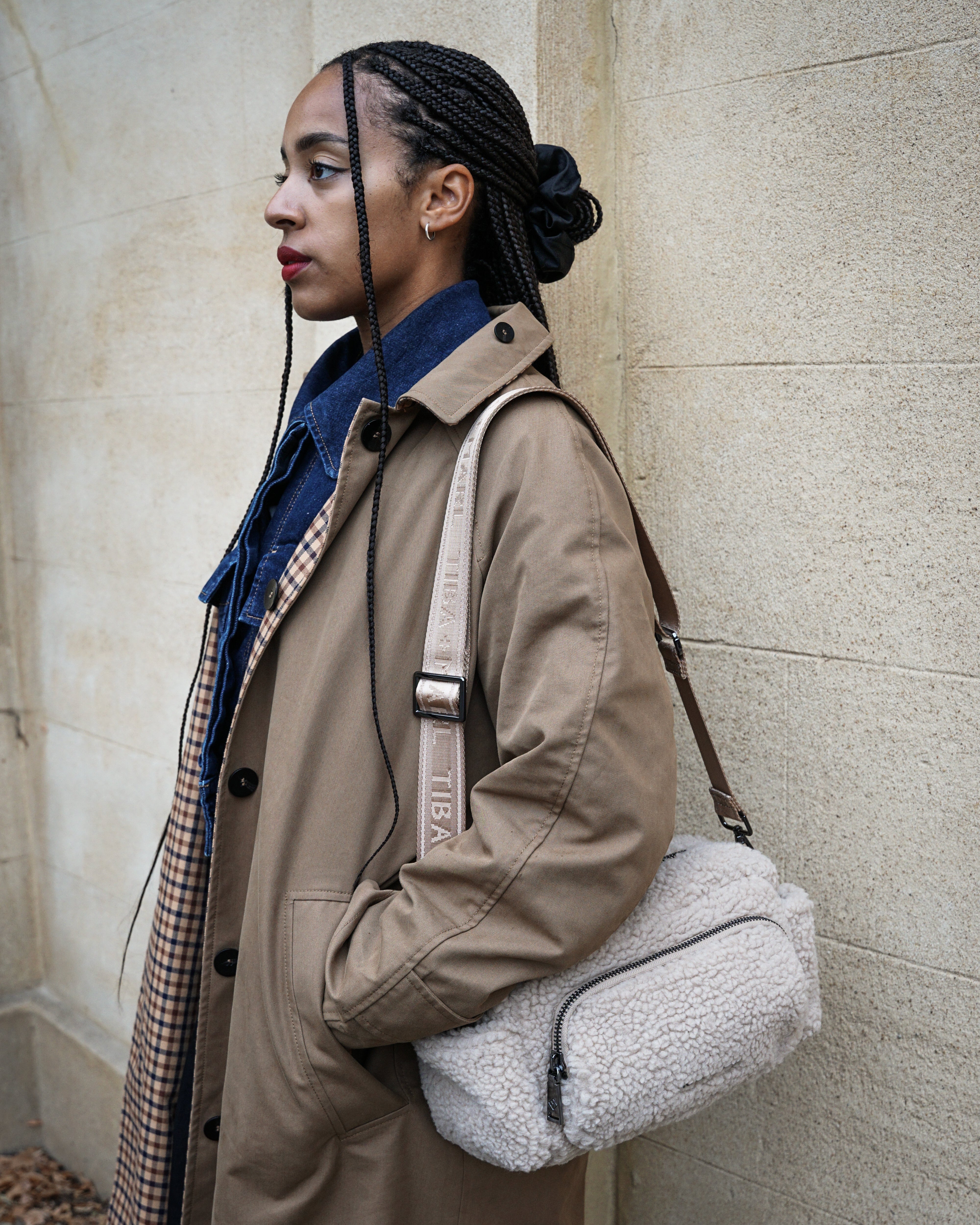 Woman in a beige trench coat holding a gray shearling bag against a stone wall.