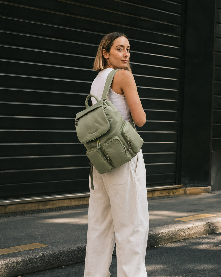 A woman wearing a white tank top and white wide-leg pants stands on a street corner in front of a black corrugated metal building, carrying an olive green backpack.