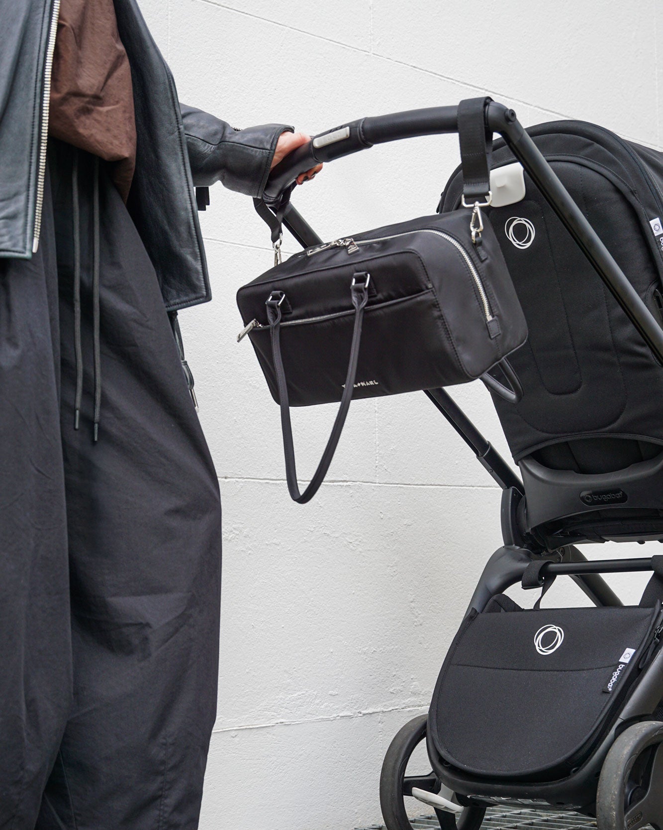 Person pushing a black stroller with a bag attached against a white wall.